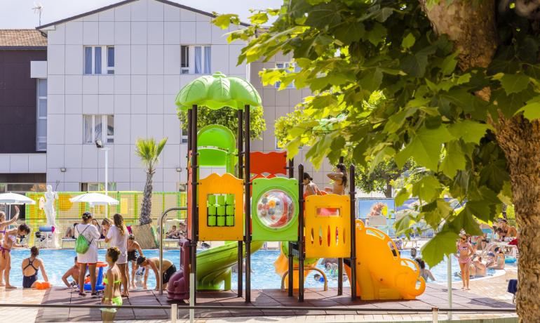 Parco giochi colorato vicino a una piscina affollata in una giornata di sole.