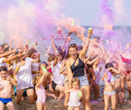 Persone festeggiano in spiaggia lanciando polvere colorata, creando un'atmosfera vivace e gioiosa.