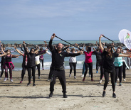 Gruppo energico si allena sulla spiaggia, sollevando bastoni, mare e cielo azzurro sullo sfondo.