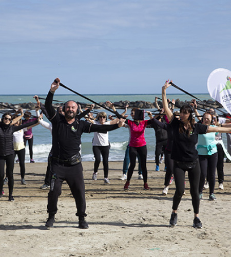 Gruppo energico si allena sulla spiaggia, sollevando bastoni, mare e cielo azzurro sullo sfondo.