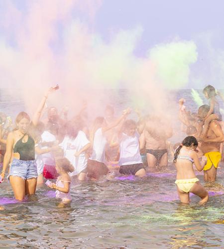 Gruppo festeggia in spiaggia lanciando polveri colorate sotto un cielo limpido.