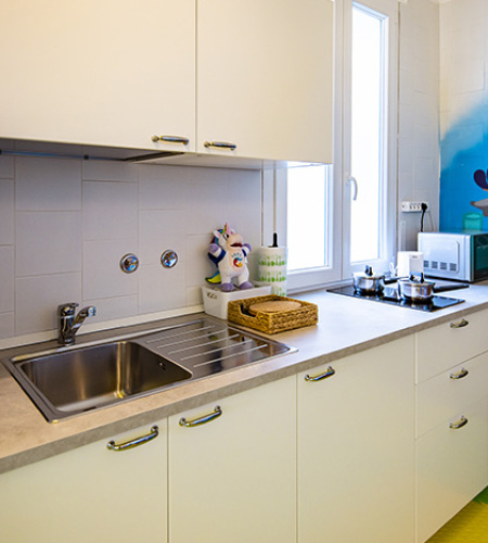 Famiglia in cucina, madre prepara, bambino sorride, altra donna dà da mangiare al piccolo.