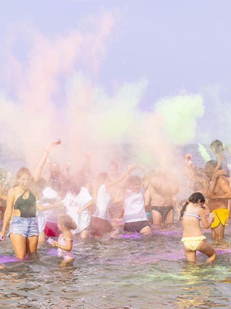 Gruppo festeggia in spiaggia lanciando polveri colorate sotto un cielo limpido.