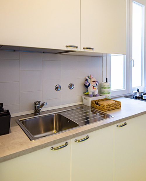 Famiglia in cucina, madre prepara, bambino sorride, altra donna dà da mangiare al piccolo.